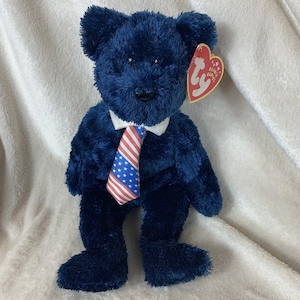 May include: A blue plush teddy bear wearing a white collared shirt and a red, white, and blue tie. The bear has a Ty Beanie Babies tag attached to its ear.