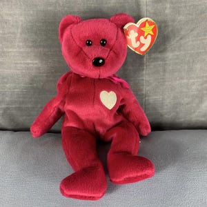 May include: A plush red beanie baby bear with a white heart on its chest. The bear has black eyes and a black nose. The bear is sitting on a grey surface. The bear has a Ty tag attached to its ear.