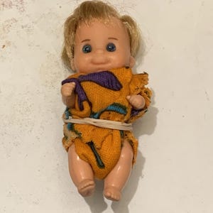 May include: A small plastic doll with blonde hair and blue eyes. The doll is wearing an orange and purple patterned outfit, secured with a rubber band. The doll's skin is a light peach color.