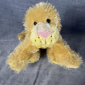 May include: A plush lion toy with shaggy, golden-yellow synthetic fur. The stuffed animal features a pink nose, black plastic eyes, and white thread whiskers. It has a rounded muzzle with small black dots representing spots on the snout.