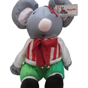 May include: A soft toy mouse with large grey ears, a white nose, and a red bow. The mouse is dressed in a red and white striped shirt, green trousers, and black shoes. A tag reading "Huggables" is attached.