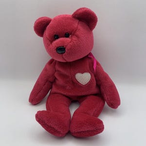 May include: A burgundy-coloured teddy bear with a white heart-shaped patch on its chest. The bear has a black nose, small black eyes, and a pink ribbon. The bear is sitting upright with its arms and legs extended.