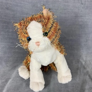 Peut inclure: Un chat en peluche avec un visage et des pattes blancs, et une fourrure rayée orange et marron. Le chat a des yeux bleus, un nez rose et des moustaches blanches. Le jouet est assis droit sur une surface grise.