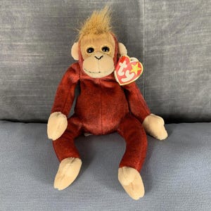 May include: A red plush monkey with a brown face and a brown tuft of hair on its head. The monkey is sitting on a grey surface and has a Ty tag attached to its arm.