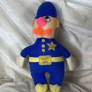 May include: A blue plush toy police officer with a pink nose, orange mustache, and a yellow star badge that says "Officer Clancy". The toy is wearing a blue uniform with a yellow belt that says "Police Dept."