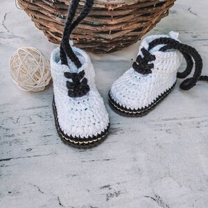 Puede incluir: Un par de botines de beb&eacute; de ganchillo blanco hechos a mano con cordones y suelas negros. Los botines se exhiben sobre una superficie blanca, con una cesta tejida y una bola decorativa en el fondo. Un accesorio encantador y acogedor.