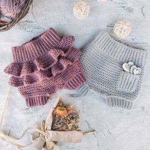 May include: Two crocheted baby bloomers, one in a light grey and one in a deep purple with ruffles. The bloomers are on a white textured surface with a bouquet of dried flowers.