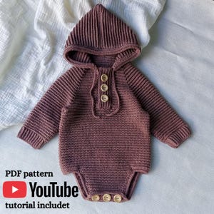 May include: A dark mauve long-sleeve hooded romper featuring a ribbed knit texture, a front placket with three light brown wooden buttons, and three matching wooden buttons at the bottom closure. The garment is displayed on a white fabric background.