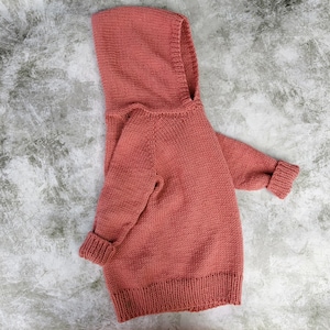 May include: A soft, knitted hooded sweater in a dusty rose color. The sweater has a relaxed fit and a long, flowing silhouette.