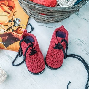 Puede incluir: Un par de zapatos de beb&eacute; de ganchillo rojo hechos a mano con cordones negros. Los zapatos tienen un estilo Oxford cl&aacute;sico, con una suela blanca y negra. Los zapatos se exhiben junto a un libro y una cesta con hilo.