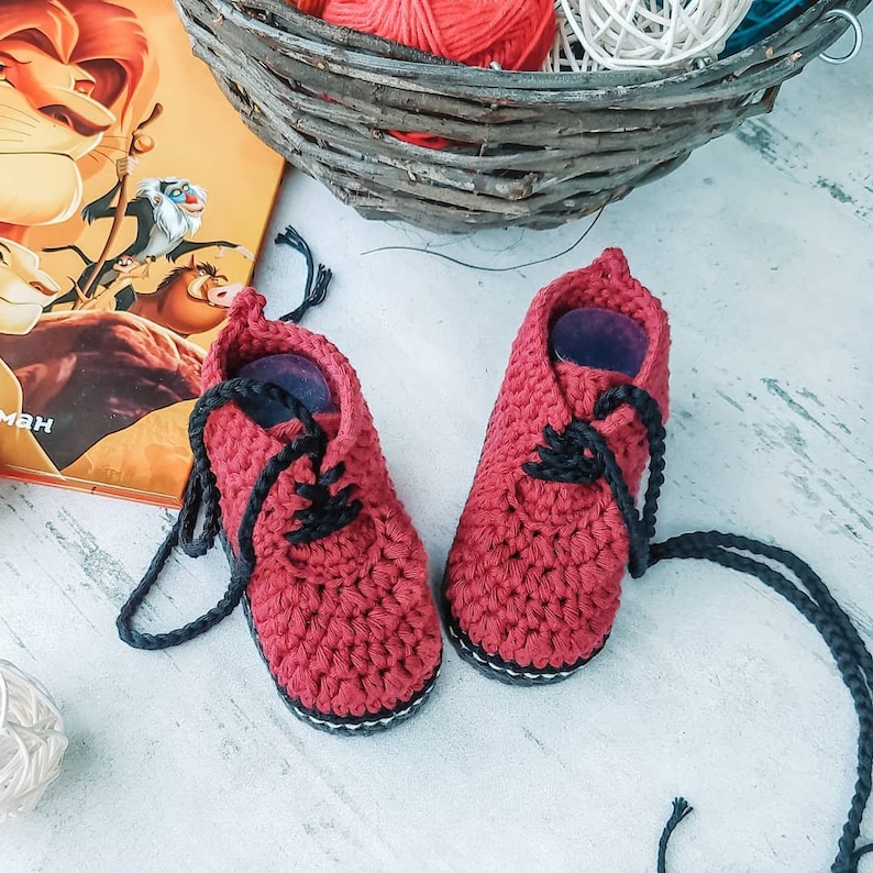 Puede incluir: Un par de zapatos de beb&eacute; de ganchillo rojo hechos a mano con cordones negros. Los zapatos tienen un estilo Oxford cl&aacute;sico, con una suela blanca y negra. Los zapatos se exhiben junto a un libro y una cesta con hilo.
