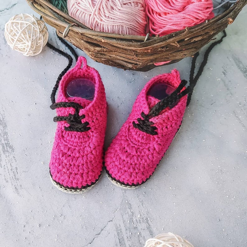 Puede incluir: Un par de zapatos de beb&eacute; de ganchillo rosa brillante hechos a mano con cordones negros. Los zapatos tienen una suela blanca y negra. El fondo incluye una cesta con hilo rosa y una bola de hilo blanco.