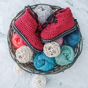Puede incluir: Un par de botines de beb&eacute; rojos hechos a mano con ganchillo, con cordones negros y un ribete gris y blanco, descansando en una cesta r&uacute;stica con varias madejas de hilo de colores y esferas decorativas. Los botines son un art&iacute;culo encantador.