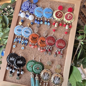 Puede incluir: Una colección de pendientes hechos a mano exhibidos en un corcho. Los pendientes están hechos de tapas de botellas, con varias marcas y diseños, con detalles de cuentas y borlas. La paleta de colores incluye azul, rojo, naranja, verde y dorado.