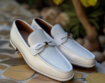 May include: A pair of white leather loafers with a textured pattern. The shoes have a bow detail and a small heel. The loafers are displayed on a stone surface, with a blurred background of greenery.
