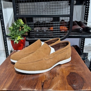 May include: A pair of brown suede slip-on shoes with white soles. The shoes have a classic design with a stitched upper and a rounded toe. The shoes are displayed on a wooden surface.