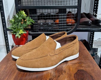 May include: A pair of brown suede slip-on shoes with white soles. The shoes have a classic design with a stitched upper and a rounded toe. The shoes are displayed on a wooden surface.