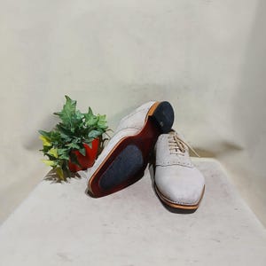 May include: A pair of light gray suede leather shoes with white laces. The shoes have a brogue design and a rounded toe.