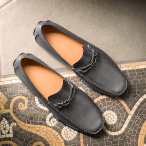 May include: A pair of black leather loafers with a braided leather detail on the vamp. The loafers have a brown leather lining and a rubber sole.