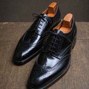 K&ouml;nnte beinhalten: Ein Paar schwarze Lederschuhe mit Brogue-Muster und Fl&uuml;gelkappe. Die Schuhe haben eine polierte Oberfl&auml;che und sind geschn&uuml;rt.