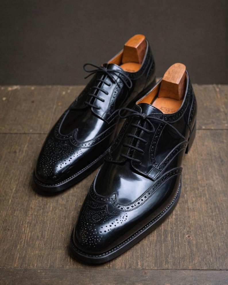 K&ouml;nnte beinhalten: Ein Paar schwarze Lederschuhe mit Brogue-Muster und Fl&uuml;gelkappe. Die Schuhe haben eine polierte Oberfl&auml;che und sind geschn&uuml;rt.