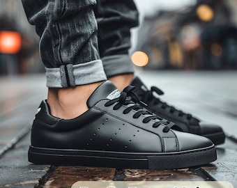 May include: Black leather low-top sneakers with white laces and a perforated design. The sneakers are worn by a person wearing dark denim jeans.