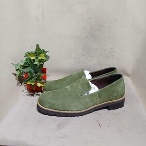 May include: A pair of olive green suede slip-on shoes with a rounded toe and a thick, dark brown sole. The shoes have a light brown trim around the sole. The shoes are new and have white paper inside. A small potted plant is in the background.