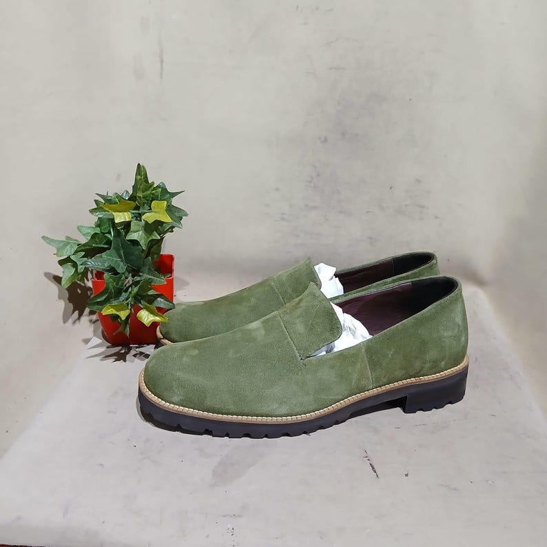 May include: A pair of olive green suede slip-on shoes with a rounded toe and a thick, dark brown sole. The shoes have a light brown trim around the sole. The shoes are new and have white paper inside. A small potted plant is in the background.