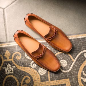 May include: A pair of brown leather loafers with a braided leather strap detail. The loafers are on a tiled floor.