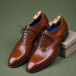 May include: A pair of brown leather lace-up shoes with a wingtip design. The shoes have a polished finish and are sitting on a wooden surface.