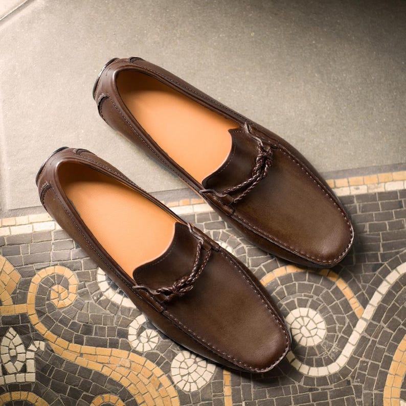 May include: A pair of brown leather loafers with a braided leather detail on the front. The loafers are on a patterned tile floor.