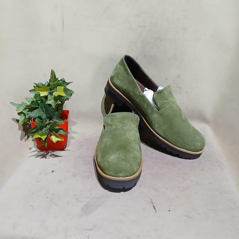 May include: A pair of olive green suede slip-on shoes with a rounded toe and a dark brown sole. The shoes are displayed on a neutral surface, with a small potted plant in the background.