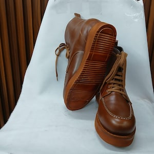 Könnte beinhalten: Ein Paar braune Lederstiefel mit passenden Schnürsenkeln und Sohlen. Die Stiefel haben ein klassisches Design mit sichtbaren Nähten und einer robusten, strukturierten Sohle. Die Stiefel sind angewinkelt und zeigen die Seiten- und Sohlendetails.