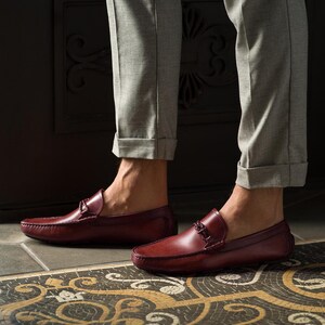 May include: A pair of burgundy leather loafers with a buckle detail. The loafers are worn by a person wearing grey pants.