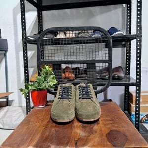 Può includere: Un paio di scarpe da ginnastica in pelle scamosciata verde oliva con lacci neri. Le scarpe sono su una superficie di legno.