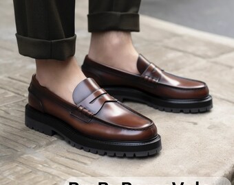 May include: A pair of brown leather loafers with a thick black sole. The loafers have a penny strap detail and are worn with dark brown pants.