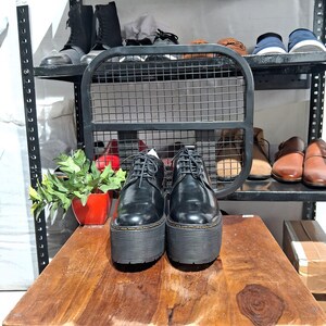May include: A pair of black platform shoes with thick soles and lace-up closures. The shoes are displayed on a wooden surface, with other shoes visible on shelves in the background. The shoes have a glossy finish.
