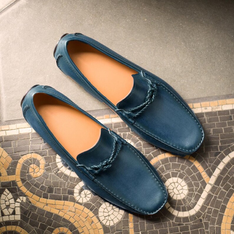 May include: A pair of blue leather loafers with a braided leather detail on the front. The loafers are on a tiled floor.