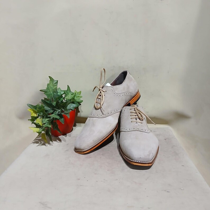May include: A pair of light gray suede leather lace-up shoes with a wingtip design. The shoes have a brown leather sole and a white lace.