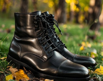 May include: A pair of black leather lace-up boots with a cap toe design. The boots are sitting on a bed of green grass and fall leaves.