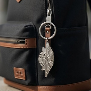 Può includere: Zaino nero con dettagli in pelle marrone e portachiavi argentato. Lo zaino presenta una tasca frontale con cerniera e una piccola toppa in pelle con il nome del marchio. Il portachiavi ha un design ad ala.