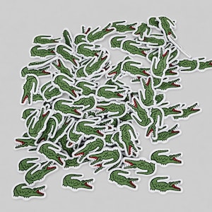 Peut inclure: Une collection de nombreuses petites pièces brodées en forme de crocodile. Chaque pièce présente un crocodile vert avec un contour blanc, une gueule ouverte rouge et des dents blanches visibles. Les pièces sont éparpillées en tas.