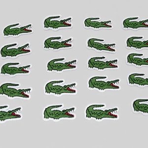 Pode incluir: Uma coleção de dezanove emblemas bordados termocolantes, cada um apresentando um crocodilo verde com boca vermelha e dentes brancos, dispostos num padrão de grelha sobre um fundo cinzento claro liso, exibindo costuras detalhadas.