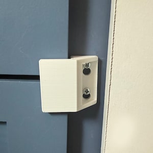 May include: A white, L-shaped bracket with two screws installed. The bracket is mounted on a blue surface, with a white surface visible on the right side of the image. The bracket appears to be made of plastic.