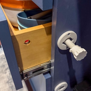 May include: Open wooden drawer with metal slides, revealing bowls and utensils. The drawer is part of a navy blue cabinet. A white, round cabinet door lock is visible on the right side of the cabinet.