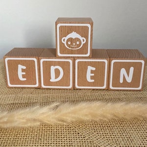 Peut inclure: Des blocs en bois orthographiant le nom "EDEN" avec un bloc de visage de singe sur le dessus. Les blocs sont brun clair avec des lettres blanches et une bordure blanche.