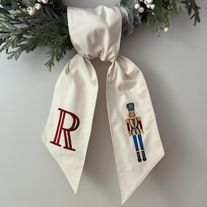 Puede incluir: Un lazo decorativo color crema con la letra "R" roja bordada en un extremo y una figura de cascanueces en el otro. El lazo cuelga de una corona verde y blanca.