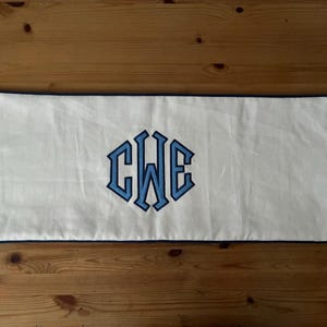 May include: A rectangular white pillow with a blue border. The center features a blue monogram of the letters C, W, and E. The pillow is placed on a wooden surface.