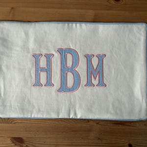 May include: Rectangular white pillow with blue trim and embroidered initials HBM in light blue with pink outlines. The pillow is made of a textured fabric and rests on a wooden surface.