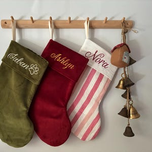 以下が含まれることがあります： 木製のラックに吊るされた3つのクリスマスストッキング。1つはオリーブグリーンで、白で「Talan」と足跡が刺繍されています。2つ目は赤で「Ashlyn」が金で刺繍されています。3つ目は白とピンクのストライプで、「Nora」が赤で刺繍されています。